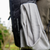 Una persona lleva una mochila Manquehue negra de 28 litros con una camisa de rayas gris claro y blancas encima. El fondo muestra una escena borrosa al aire libre con vegetación.